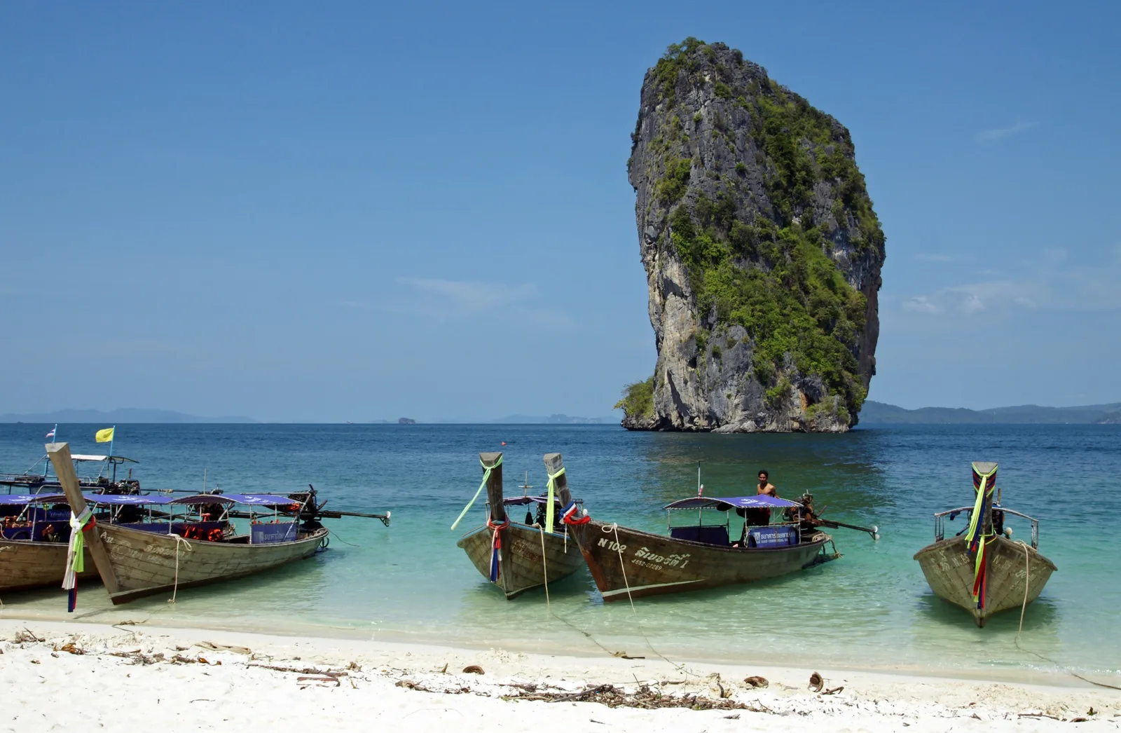 Longtail boats moored on a white-sand beach with a tall limestone island behind, Krabi